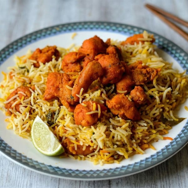 Chicken biryani with orange chicken pieces and a lime wedge on a white plate with blue trim.