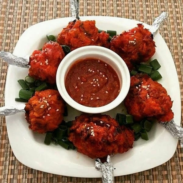 Six red chicken lollipops with foil-wrapped bones, green onions, and a dipping sauce on a white plate.